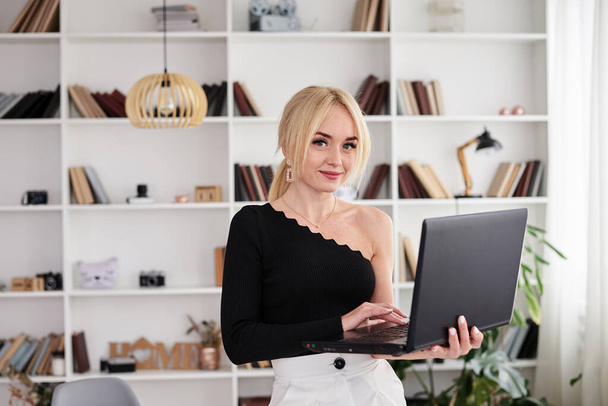 Young blond woman, wearing black top and white pants, standing by the table, holding laptop. Office manager at her workplace, preparing for presentation. Female working on computer in light office. - Фото, изображение