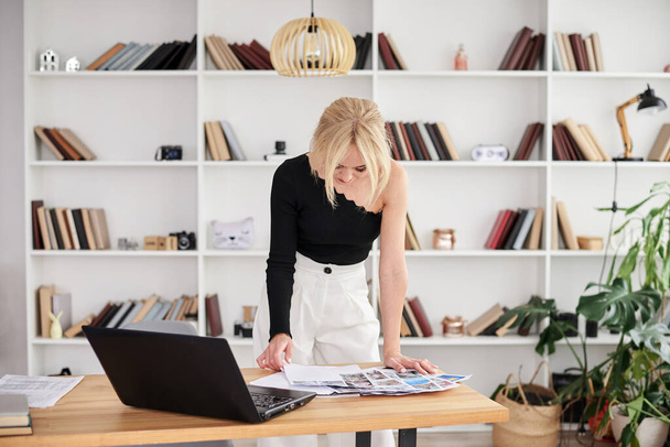 Young blond woman, wearing black top and white pants, standing by table, working on laptop. Office manager at her workplace, preparing for presentation. Female working in light office in company. - Foto, immagini