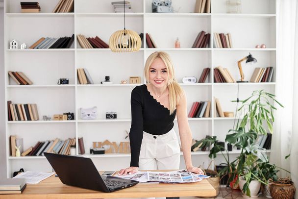 Young blond woman, wearing black top and white pants, standing by table, working on laptop. Office manager at her workplace, preparing for presentation. Female working in light office in company. - Foto, immagini