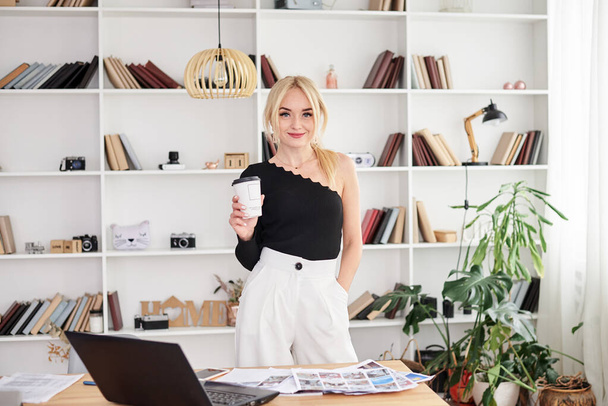 Young blond woman, wearing black top and white pants, standing by table with laptop, holding paper coffee cup. Office manager at her workplace. Female working in light office in company. - Foto, Imagen