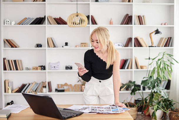 Young blond woman, wearing black top and white pants, standing by table, talking on phone. Office manager at her workplace, preparing for presentation. Female working in light office in company. - Photo, image