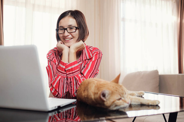 bisnesnainen punaisessa paidassa punapäisen kissan kanssa istuu toimistossa pöydässä ja katselee avointa kannettavaa tietokonetta. Nainen kommunikoi videoneuvottelun kautta kissan ollessa pöydällä - Valokuva, kuva