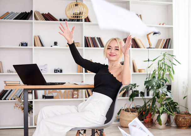 Young blond woman, wearing black top and white pants, sitting on modern grey chair, throwing papers in the air.Office manager at her workplace. Female working in company. Busy workday. - Foto, Imagem