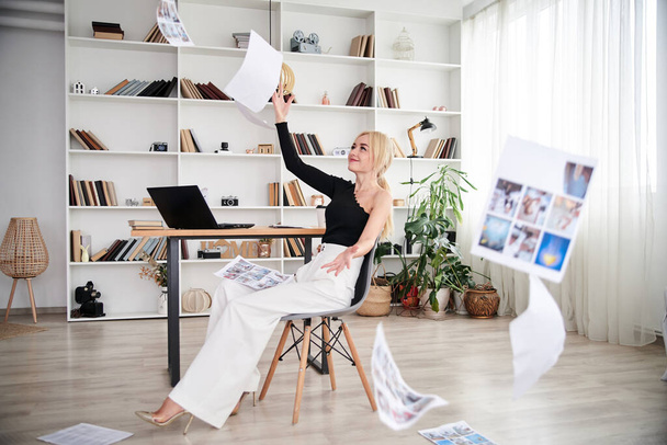 Young blond woman, wearing black top and white pants, sitting on modern grey chair, throwing papers in the air.Office manager at her workplace. Female working in company. Busy workday. - Valokuva, kuva
