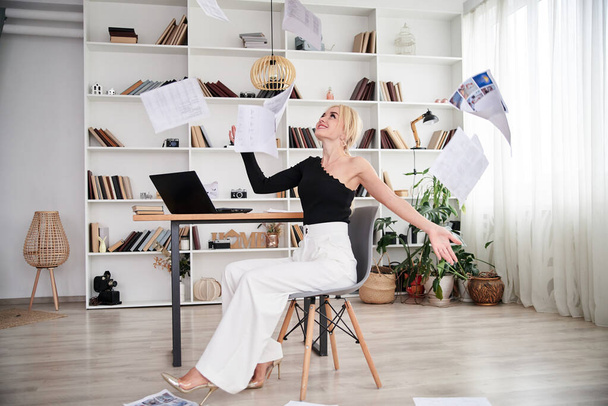 Young blond woman, wearing black top and white pants, sitting on modern grey chair, throwing papers in the air.Office manager at her workplace. Female working in company. Busy workday. - Photo, image