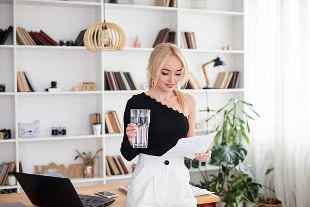Young blond woman, wearing black top, sitting on office table, holding huge glass of water and paper document, smiling.Manager at workplace. Work process in company.Healthy drink consumption concept. - Zdjęcie, obraz