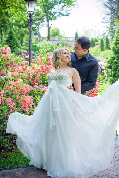 Mooie blonde bruid met een prachtige vormen in de tuin tussen de bloemen in een lange lichte jurk. Bruid en bruidegom op de wandeling - Foto, afbeelding