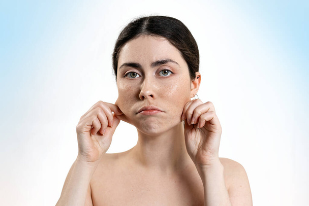 Portrait of a distressed caucasian woman, pulling the skin on her cheeks with her hands. Light Blue background. Concept of skin care. - Photo, Image