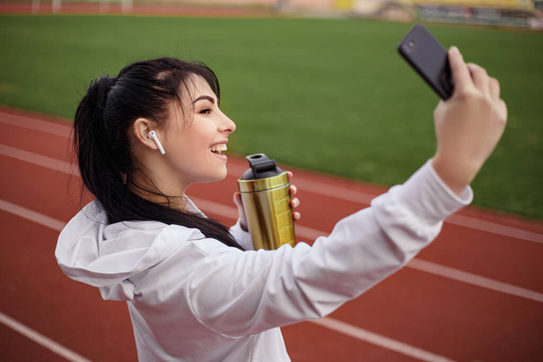 Young athletic girl drink bottle shaker fresh water protein after productive fitness workout on running track treadmill. Female sporty model making selfie smiling smartphone hand gold. - Photo, Image