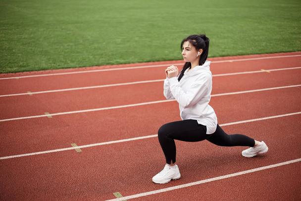 Girl stretches, prepares her body and muscles for a productive fitness workout. Flexible female dressed sports suit sporty model on the city stadium. Image with copy space. Single leg squats - Valokuva, kuva