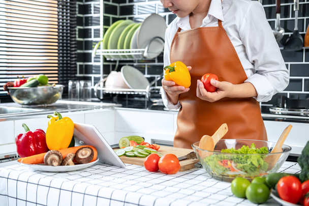 Asian housewife holding bell pepper and tomato in hands during watching tutorial cooking class online on tablet for preparing to cooking healthy meal in the kitchen at home - Φωτογραφία, εικόνα