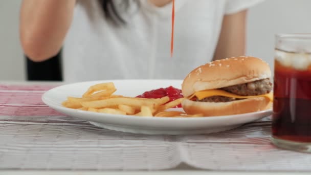Close-up shot richt zich op ketchup in rode fles die werd gegoten door de vrouw op een witte schotel op tafelkleed, om te eten met junk food, dat is ongezond, zoals hamburgers, frietjes, en cola. - Video