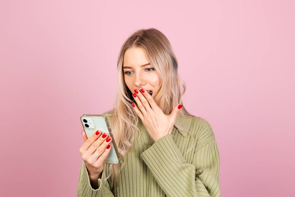 Bella donna europea in maglione casual a maglia su sfondo rosa isolato sguardo felice al telefono con volto sorpreso stupito - Foto, immagini