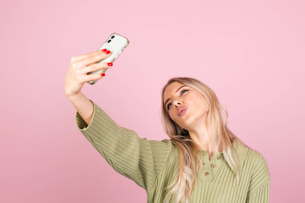 Mooie europese vrouw in casual gebreide trui op roze achtergrond glimlachen en kijken naar de camera terwijl het nemen van selfie foto stuur lucht kus - Foto, afbeelding