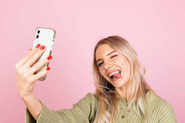 Jolie femme européenne en pull tricoté décontracté sur fond rose prenant selfie photo sur téléphone fait grimaces drôles montre la langue positive - Photo, image