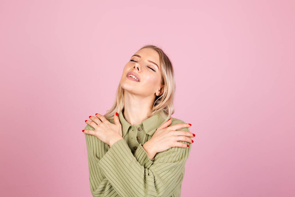 Pretty european woman in casual knitted sweater on pink background self care hugging herself with smile and closed eyes - Fotografie, Obrázek