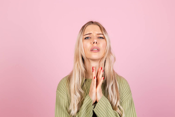 Hübsche Europäerin in lässigem Pullover auf rosa Hintergrund betet mit Händen und hoffnungsvollem Gesichtsausdruck sehr emotional und besorgt - Foto, Bild