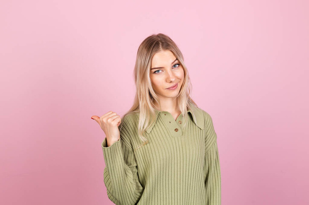 Mulher europeia bonita em camisola casual no fundo rosa feliz ponto de sorriso à esquerda com o dedo polegar - Foto, Imagem