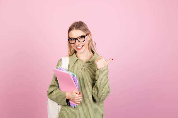 Pretty european woman in casual sweater on pink background with notebooks smiling happy pointing thumb finger right education concept - Photo, Image