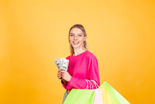 Pretty european kobieta w różowej bluzce na żółtym tle uśmiech podekscytowany trzymać fan 100 dolarów pieniędzy i torby na zakupy, podekscytowany zaskoczony uśmiechnięty szczęśliwy - Zdjęcie, obraz