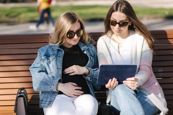 Pregnant woman with her best friend sitting in the park and looking for baby stroller using tablet. Two females in sunglasses choose clothes for baby - Fotó, kép