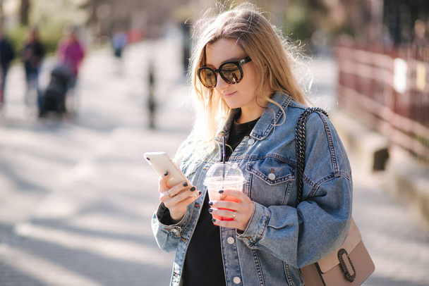 Mujer bastante prengant bebiendo limonada de fresa mientras camina al aire libre. Retrato de atractiva hembra en gafas de sol usando teléfono - Foto, Imagen