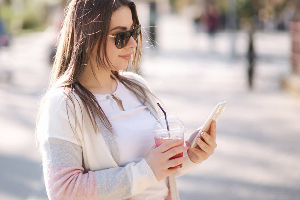Kaunis nainen käyttää puhelinta ja juo limonadia ulkona. Nuori bloggaaja viettää aikaa puistossa - Valokuva, kuva