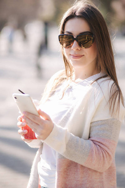 Όμορφη γυναίκα που χρησιμοποιεί το τηλέφωνο και πίνει λεμονάδα εξωτερική. Νεαρός blogger περνάει χρόνο στο πάρκο - Φωτογραφία, εικόνα