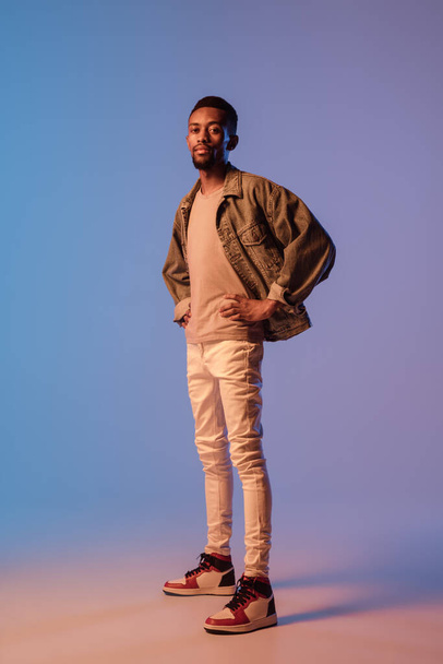 Young stylish man in modern street style outfit isolated on gradient background in neon light. African-american fashionable model in look book, musician performing. - Zdjęcie, obraz