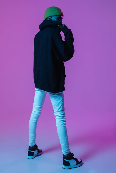 Young stylish man in modern street style outfit isolated on gradient background in neon light. African-american fashionable model in look book, musician performing. - Fotó, kép