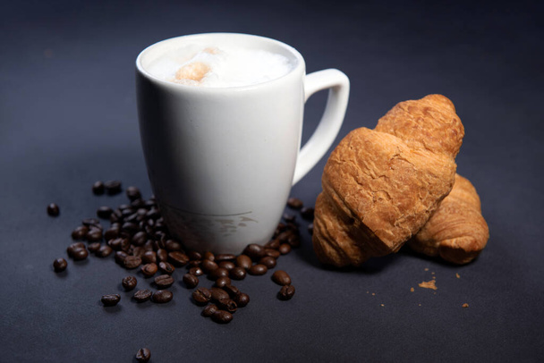 Beautiful latte macchiato with milk foam in a white cup with croissants and coffee beans nearby. Dark blue background. A cup of coffee with milk. Hot latte or cappuccino on milk with copy space - Zdjęcie, obraz