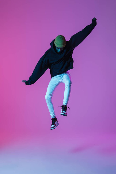 Young stylish man in modern street style outfit isolated on gradient background in neon light. African-american fashionable model in look book, musician performing. - Fotografie, Obrázek