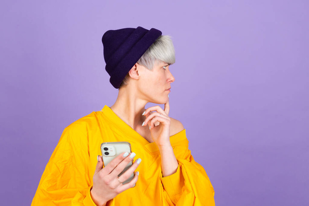 Stylish european woman on purple background thinking dreaming worry holding mobile phone - Photo, Image