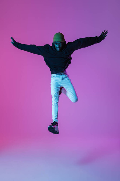 Young stylish man in modern street style outfit isolated on gradient background in neon light. African-american fashionable model in look book, musician performing. - Φωτογραφία, εικόνα