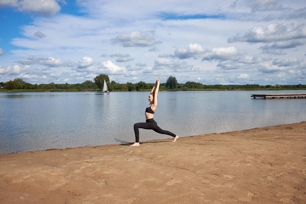 Jovem feliz vestindo sportswear preto praticando ioga na areia perto do lago ao ar livre. - Foto, Imagem