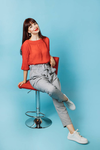 Pretty smiling Caucasian woman in stylish clothes, sitting on chair against blue background. Full length portrait of young businesslady with bright makeup, sitting on the chair - Фото, зображення
