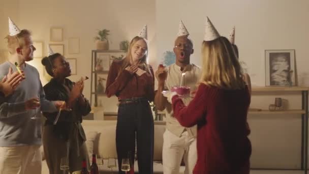 Medium shot van een groep vrienden blazen feesthoorns en klappen terwijl jonge vrouw blaast kaars op taart. Jongeren vieren thuis hun verjaardag - Video