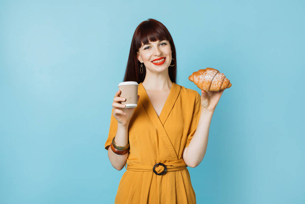 Street food, coffee break. Image of cheerful red haired young woman in trendy yellow suit, holding fresh tasty croissant and take away coffee, posing isolated over blue background. Copy space - Фото, изображение