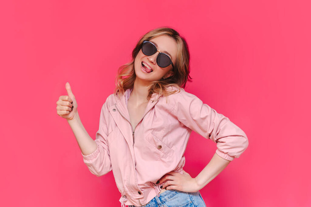 A young caucasian beautiful happy cheerful smiling brown-haired woman in a casual jacket and sunglasses shows tongue and thumb up like gesture with her hand isolated on a bright color pink background - Photo, Image