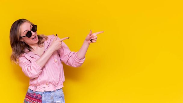 A young caucasian excited smiling  woman in a pink jacket and sunglasses indicate copy empty blank space for text or design with two fingers presenting product on bright color yellow background.Banner - Foto, imagen