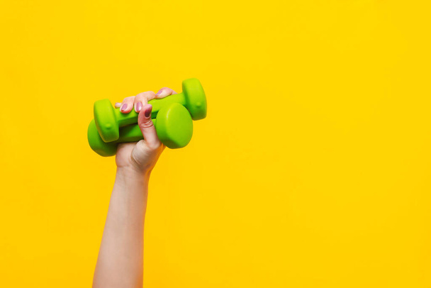 Two green dumbbells in a woman's hand isolated on yellow background. Sport equipment. Fitness concept, healthy lifestyle. Copy space for design - Foto, Imagem