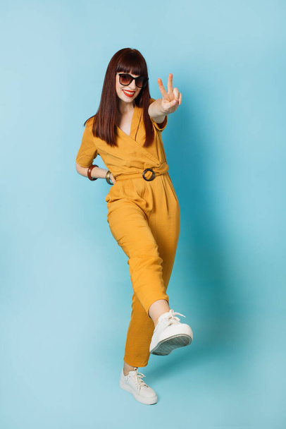 Young beautiful brunette woman wearing stylish ocher suit, smiling with happy face and showing her leg at the camera, doing victory sign over blue background. Number two, peace and victory sign. - Photo, Image