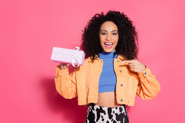 amazed african american woman in crop top and jacket pointing at present on pink - Photo, Image