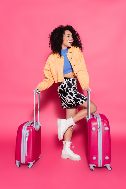 longitud completa de la elegante mujer afroamericana sonriendo mientras posando con maletas en rosa  - Foto, Imagen
