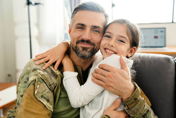 Onnellinen maskuliininen sotilaallinen mies halaa hymyilevää tytärtään istuessaan sohvalla sisätiloissa - Valokuva, kuva