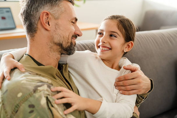 Happy masculine military man hugging his smiling daughter while sitting on sofa indoors - Foto, imagen