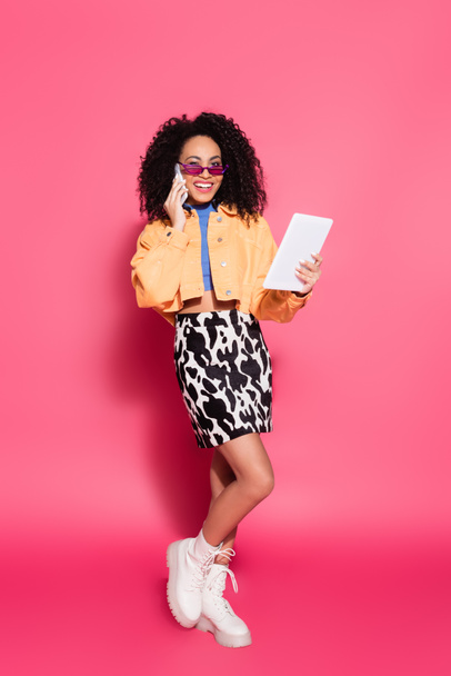 full length of happy african american woman in sunglasses talking on smartphone while holding digital tablet on pink - Фото, зображення