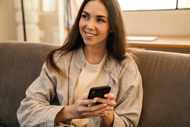 Happy young woman smiling and using smartphone while sitting on sofa indoors - Foto, Imagem