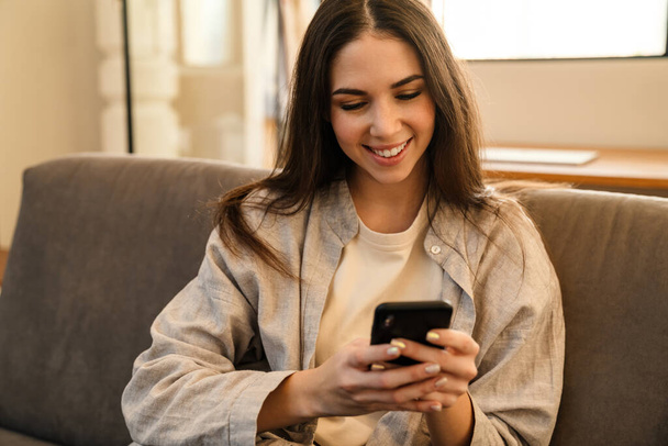 Happy young woman smiling and using smartphone while sitting on sofa indoors - Φωτογραφία, εικόνα