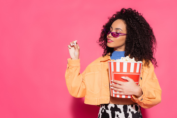 african american woman in sunglasses holding bucket and popcorn on pink  - Фото, зображення